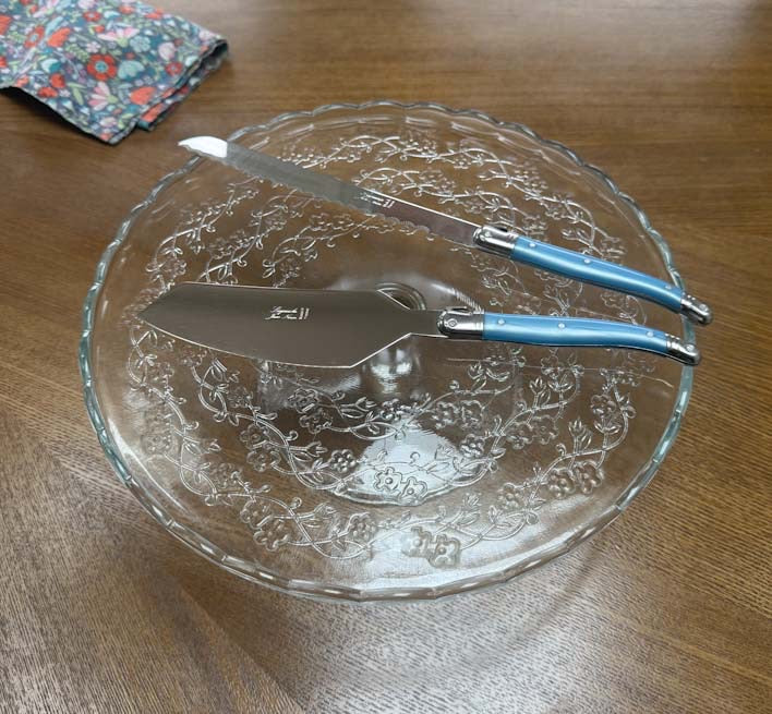 A clear glass serving platter with an embossed floral pattern sits on a wooden table, with a cake server and a bread knife featuring light blue Laguiole handles. Clementine Boutique Made in France