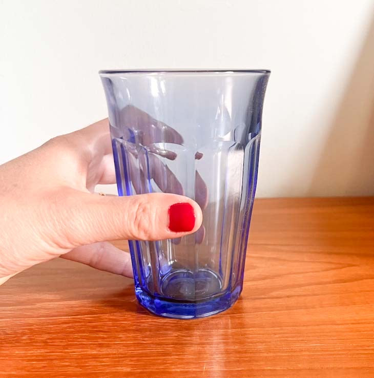 Hand holding a Duralex Marine Picardie Highball 36cl glass on a wooden surface with a white background, made in France