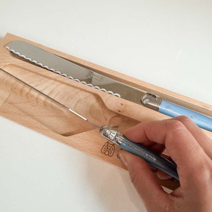 A hand holding a Laguiole small blue-handled knife with a bee detail over a wooden knife block, with a larger serrated knife resting in the block. The scene is photographed on a plain white background.