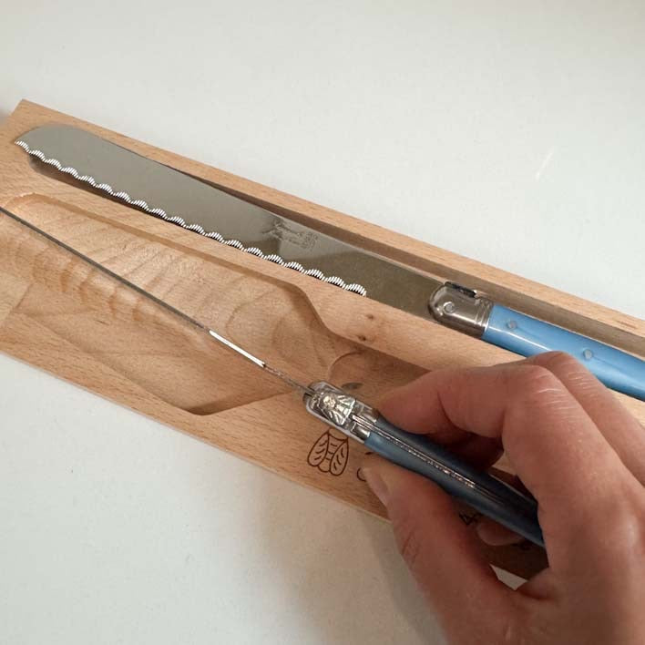 A close-up of a hand holding a blue-handled knife over a wooden knife block, with another serrated knife resting in the block. The knives have silver blades and blue handles with bee silver metal accents. Clementine Boutique Made in France