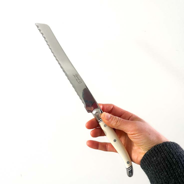 A Laguiole bread knife with an ivory handle, placed on a red basket containing a partially sliced loaf of bread and a cloth with a floral pattern.