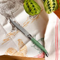 A Laguiole kitchen knife with a sage green handle with a bee emblem and a silver blade lies diagonally on a cream-colored kitchen towel printed with farm animals and red stripes. Two green plant leaves are visible in the background.