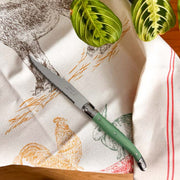 A Laguiole kitchen knife with a sage green handle with a bee emblem and a silver blade lies diagonally on a cream-colored kitchen towel printed with farm animals and red stripes. Two green plant leaves are visible in the background.