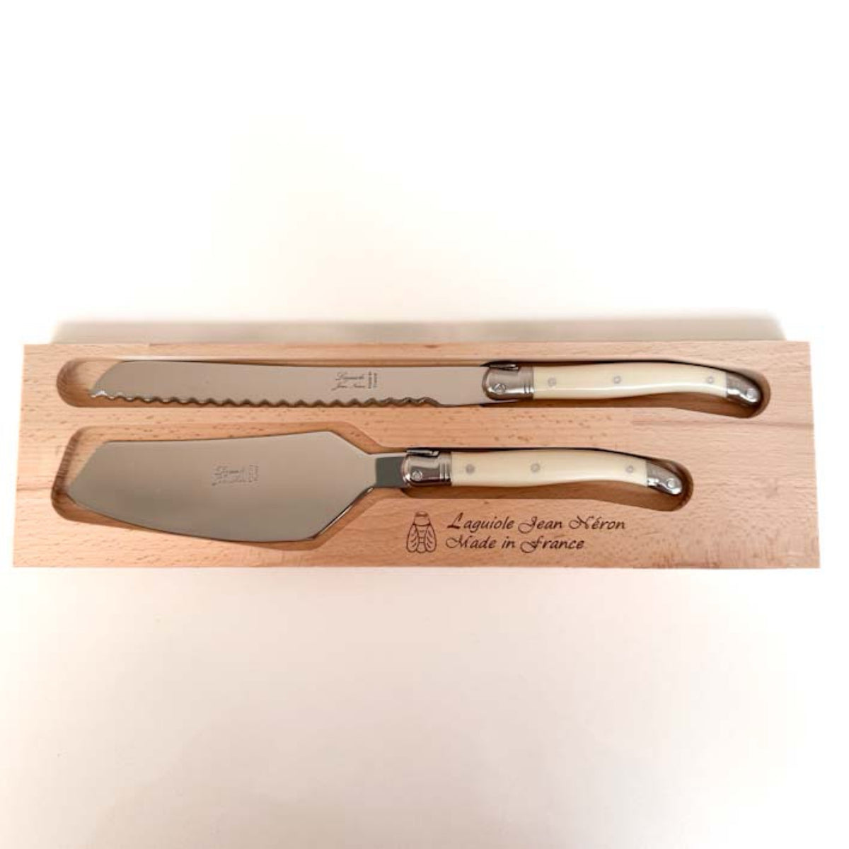 A two-piece wooden serving knife set from Laguiole Jean Neron, featuring a serrated bread knife and a cake/pie server, both with cream-colored handles and polished steel blades, presented in a engraved wooden box.