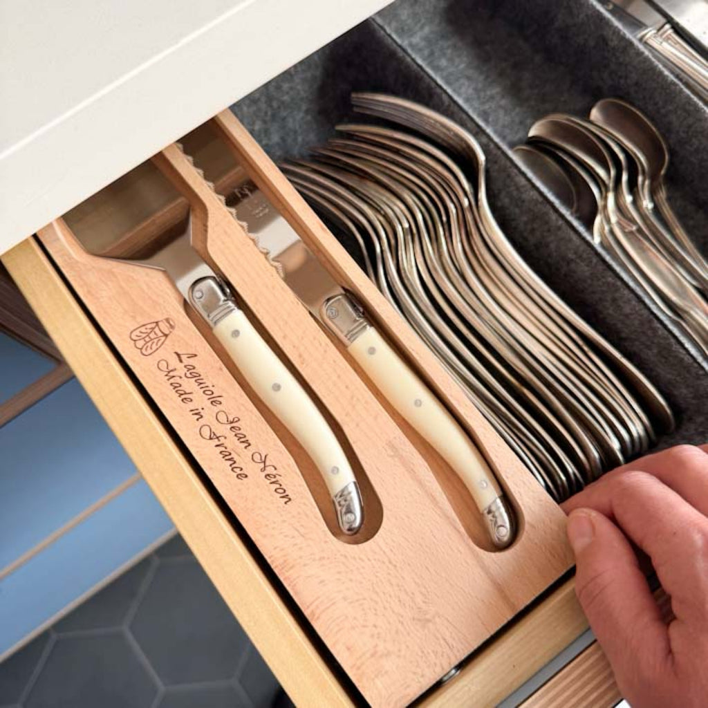 A close-up of a wooden drawer organizer holding a set of cutlery composed of a cake server and a bread knife with cream-colored handles in a wooden tray with an engraved text reading 'Laguiole Jean Neron Made in France.