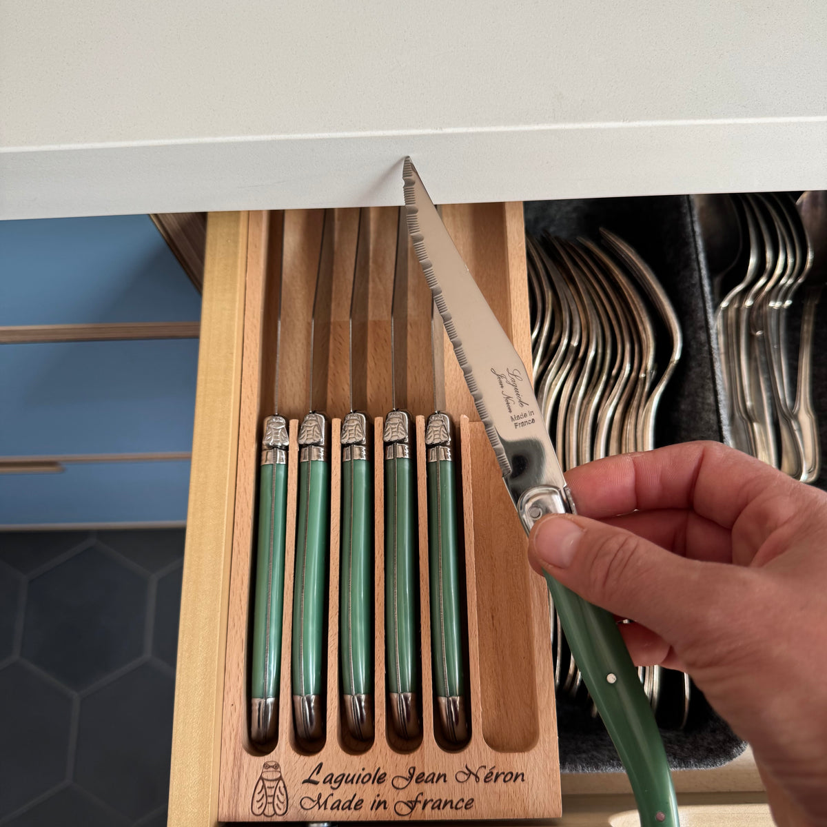 A hand holds a Laguiole Jean Néron steak knife with a green handle and serrated stainless steel blade, positioned over an open wooden drawer containing matching Laguiole Jean Néron steak knives and forks.