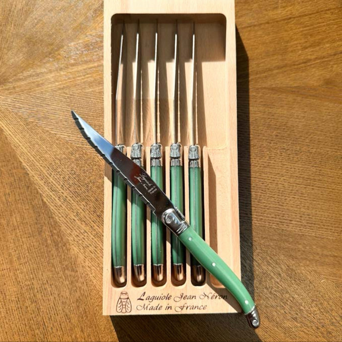 A set of five Laguiole Jean Neroon steak knives in a light wood presentation box, with one knife opened and resting diagonally across the front. The knives feature green handles with serrated stainless steel blades, set on a wooden tabletop.