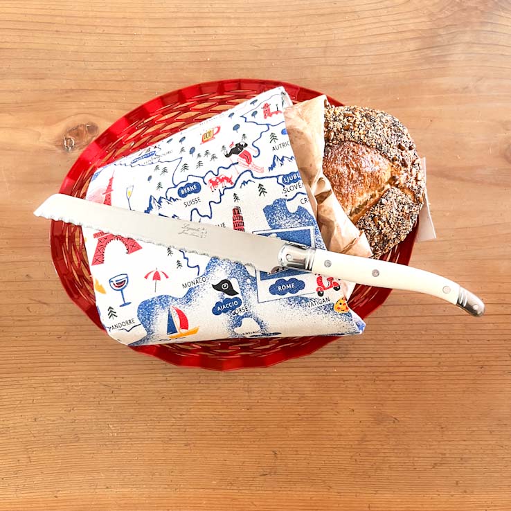 Red basket with bread, a Laguiole Bread knife, and a decorative napkin on a wooden surface